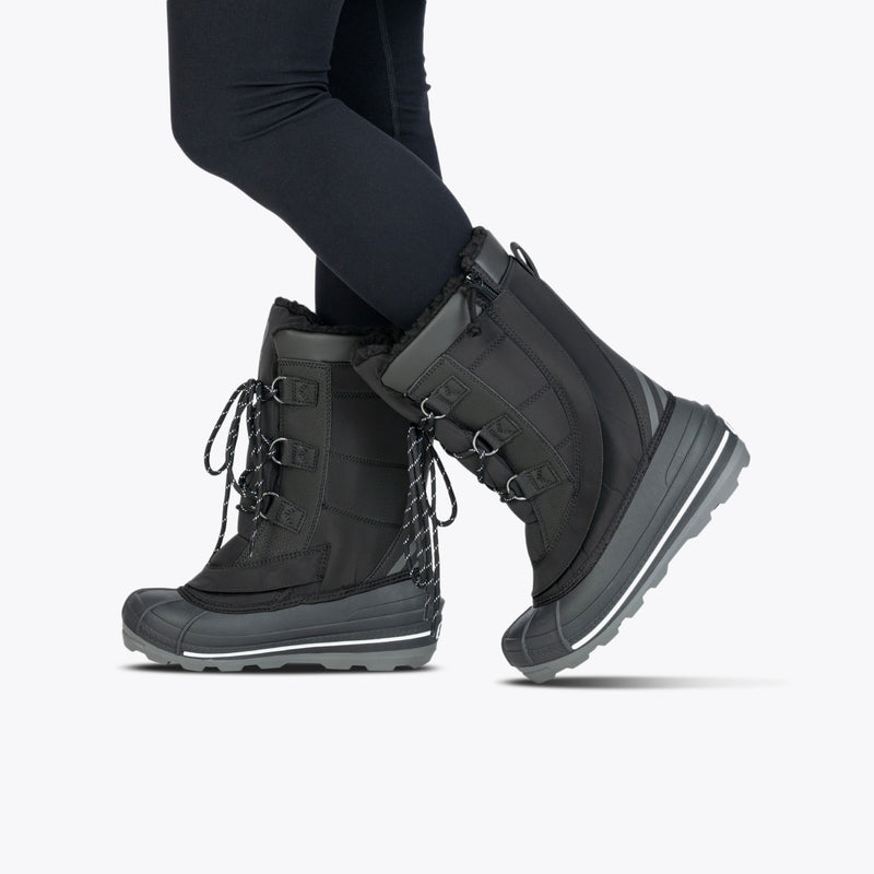 A person wearing Black BILLY Ice Boot by BILLY Footwear, featuring thick soles, fur lining, and lace-up fronts, is paired with black leggings and stands against a plain light background.