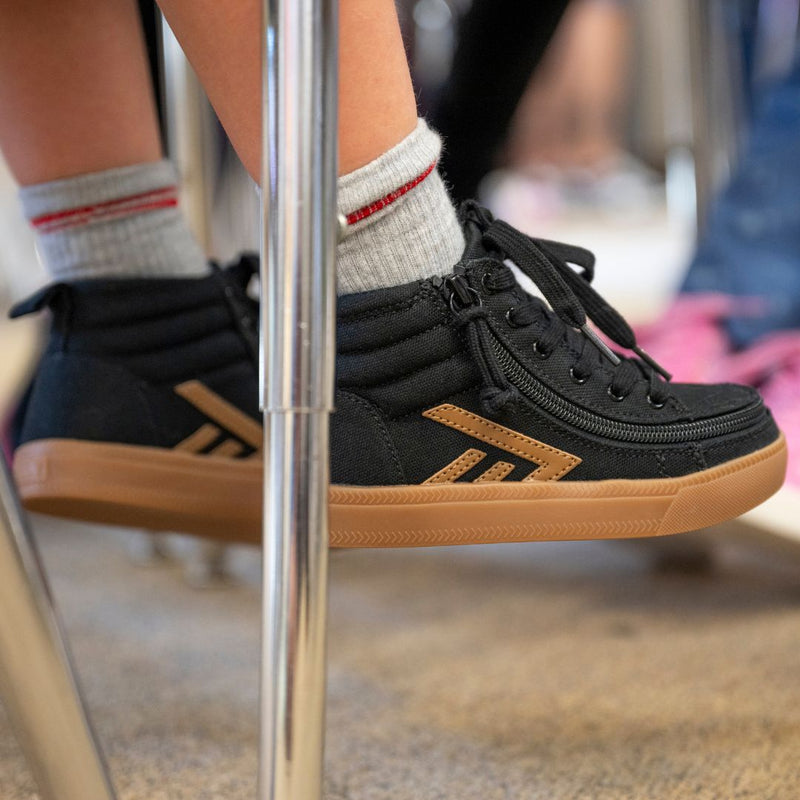 A close-up of a person’s feet in Black/Gum BILLY CS High sneakers by BILLY Footwear, paired with white socks featuring red stripes, resting on the crossbar of a metal chair in a classroom.