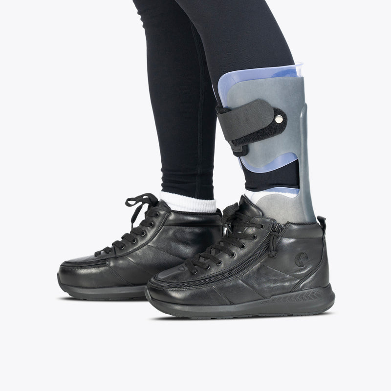 Wearing black leggings and a gray plastic AFO brace on their lower right leg, a person showcases the BILLY Footwear Black Leather BILLY Goat Classic High shoes against a plain background.