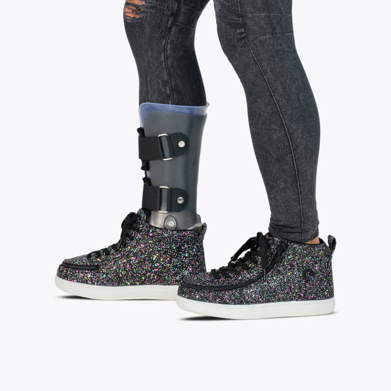 Wearing distressed black jeans, an orthotic leg brace, and BILLY Footwear’s Black Multi Glitter BILLY Classic D|R II High shoes with white soles, a person stands against a plain white background.