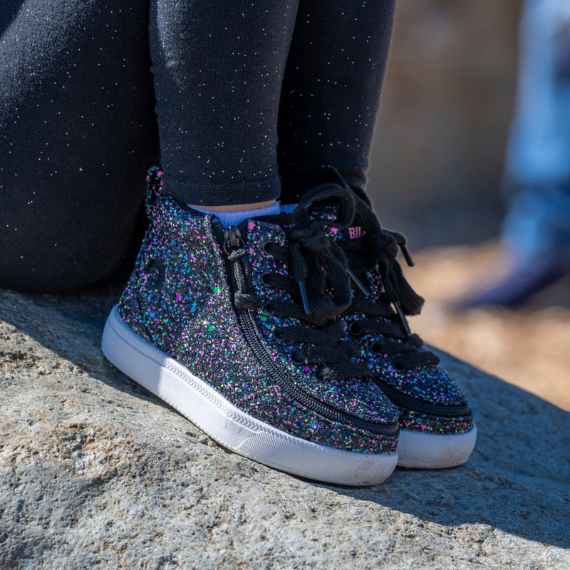 A close-up of a child’s legs wearing black leggings with white specks and BILLY Footwear’s Black Multi Glitter BILLY Classic D|R II High, AFO-friendly kids shoes for braces, sitting on a large rock outdoors.