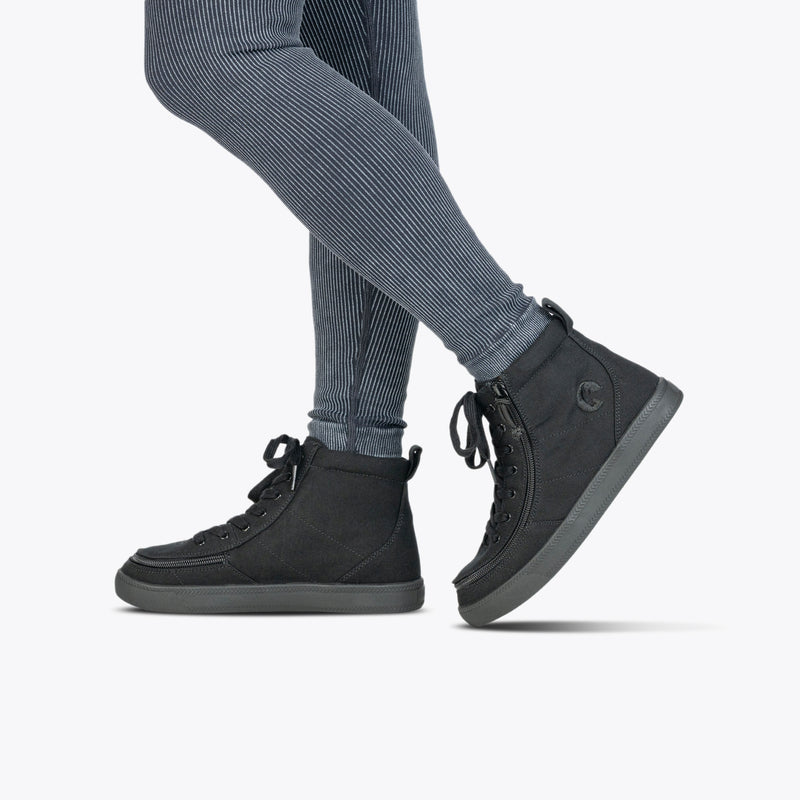 Person wearing ribbed dark gray leggings and Black to the Floor BILLY Classic Lace High sneakers by BILLY Footwear, featuring wraparound zippers and dark gray soles, stands on one foot with a plain white background.