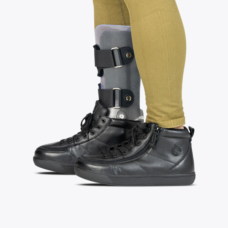 A person in yellowish pants wears BILLY Footwear's Black to the Floor Leather BILLY Classic D|R II High, AFO-friendly shoes, with a gray leg orthosis brace on their left leg. The background is plain white.