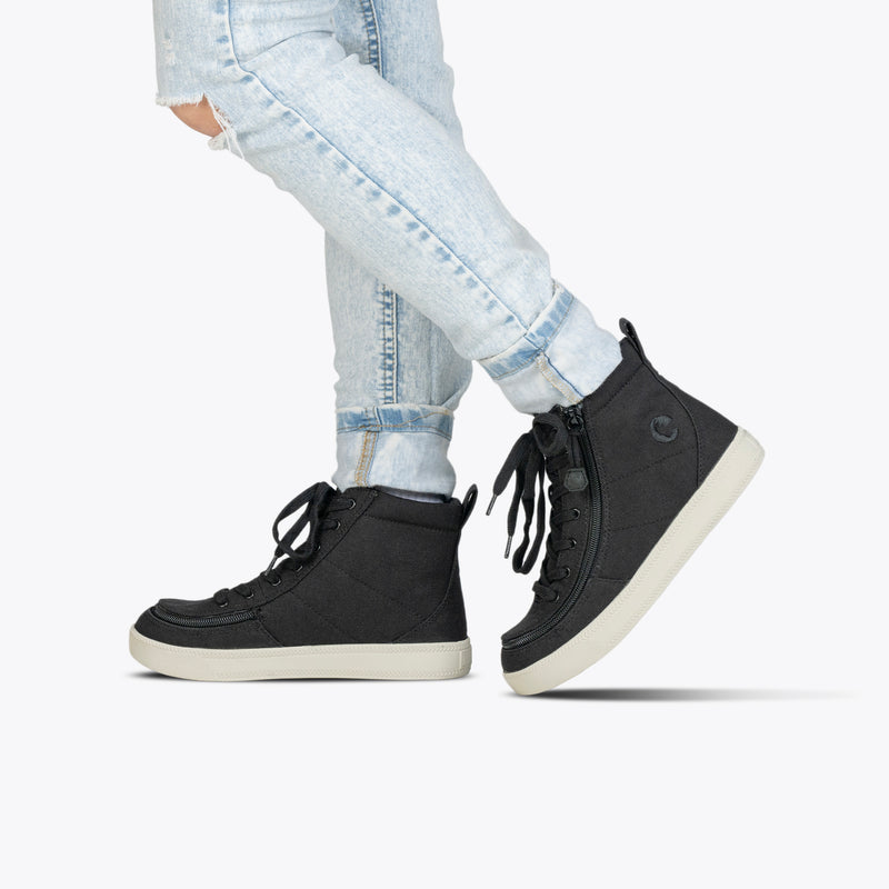 Someone stands on one foot, lifting the other slightly, in light-wash rolled-up jeans and Black/White BILLY Classic Lace High sneakers by BILLY Footwear, against a plain white background.