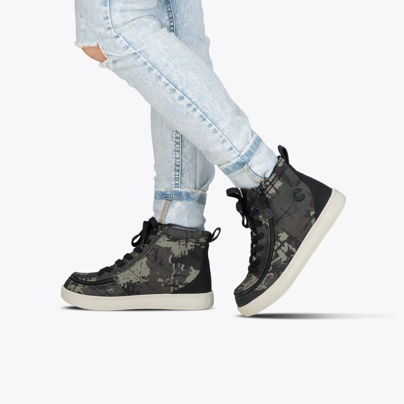 A person stands on a white background wearing light blue ripped jeans and Camo/White BILLY Classic Lace High sneakers from BILLY Footwear, featuring a grey camo pattern and signature wraparound zipper.