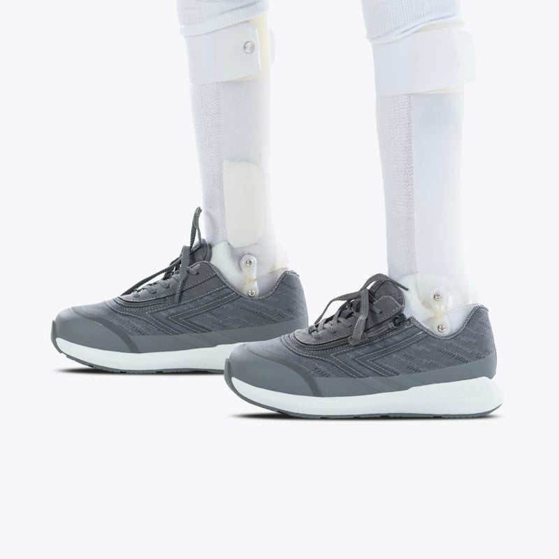 A person with white prosthetic lower legs wears Charcoal BILLY Goat shoes by BILLY Footwear, standing on a white background. The focus is on the AFO-friendly design and easy on-and-off zipper feature of the shoes and prosthetics.