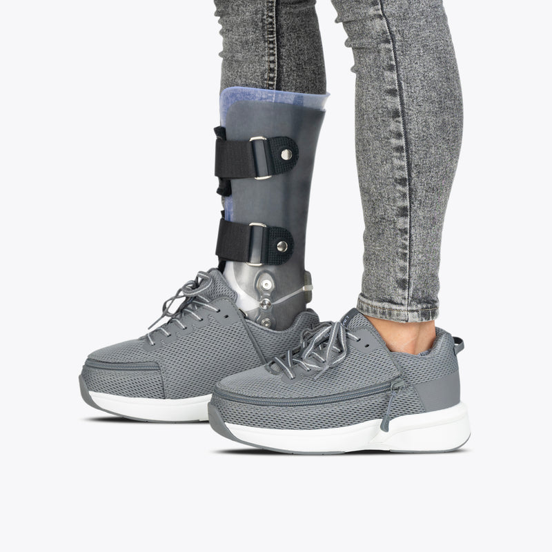 A person wearing Charcoal BILLY Limitless shoes by BILLY Footwear and jeans, with an orthotic leg brace visible on one leg, stands against a plain light background.