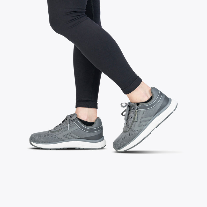 A person in black leggings and Charcoal BILLY Sport Inclusion shoes by BILLY Footwear stands against a white background, with one foot flat on the ground and the other lifted onto its toes.