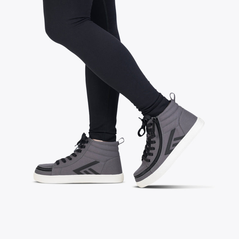 A person in black leggings and Charcoal/Black BILLY CS High sneakers by BILLY Footwear, featuring a full wrap zipper, stands on one leg with the other raised and bent at the knee against a plain white background.