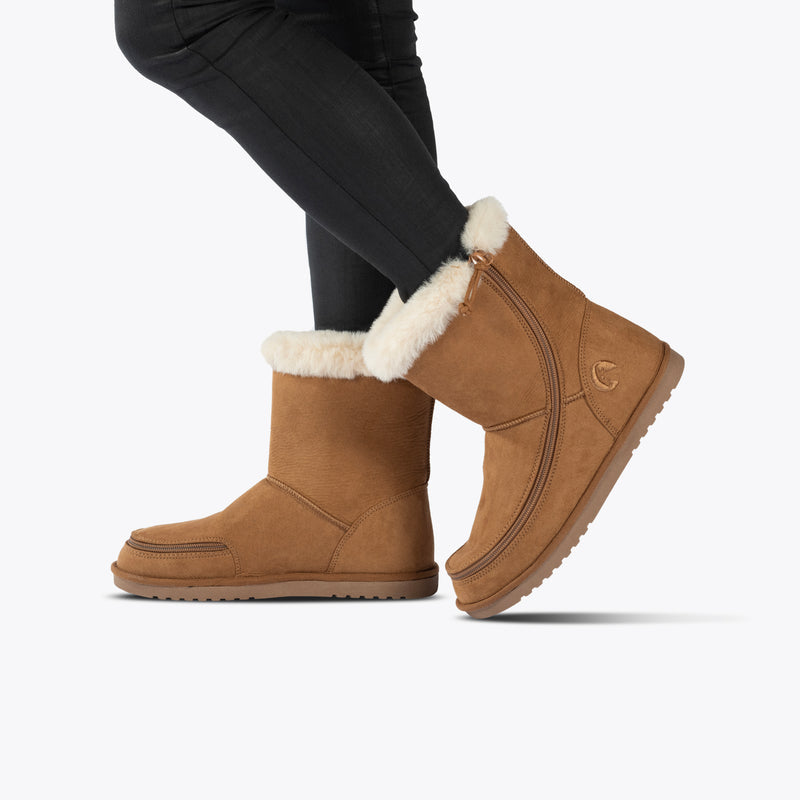 Wearing black pants and Chestnut BILLY Cozy II boots by BILLY Footwear, a person stands on one foot with the other leg slightly raised behind, posed against a plain white background.