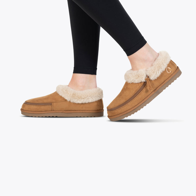 A person wears black leggings and Chestnut BILLY Cozy Slippers by BILLY Footwear, featuring a plush cream lining, thick soles, and a soft, cozy design for comfort, shown on a white background.