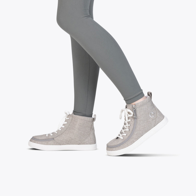 A person wearing grey leggings and BILLY Footwear’s Grey Jersey BILLY Classic Lace Highs with white soles, laces, and a wraparound zipper, walking against a plain white background.