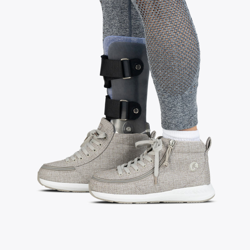 A person wearing Grey Jersey BILLY Goat Classic High shoes by BILLY Footwear, with one shoe fitted over a black and gray orthopedic leg brace, stands on a white background in gray textured leggings and white socks.