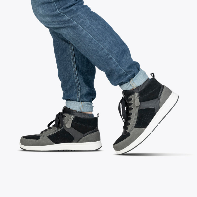A person wearing BILLY Footwear's Men's Black/Charcoal/Black BILLY Comfort DZ High sneakers and blue jeans walks with one foot slightly raised against a plain white background, highlighting these comfortable shoes.