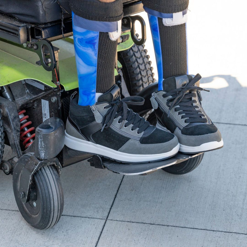 A person in blue orthotic leg braces and Men's Black/Charcoal/Black BILLY Comfort DZ High shoes by BILLY Footwear sits in a green motorized wheelchair on a sidewalk.
