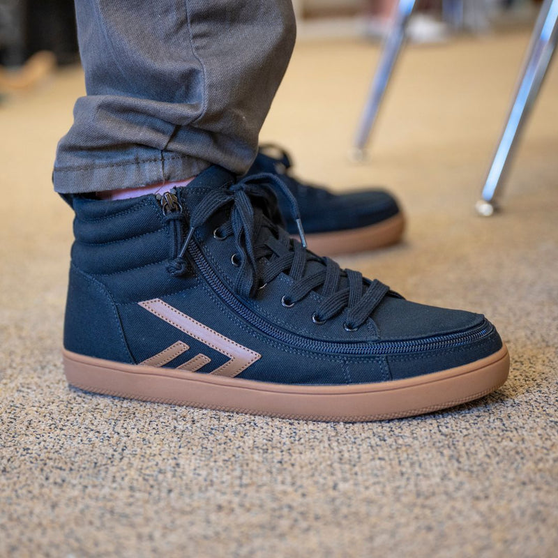 A person wearing BILLY Footwear Men's Black/Gum BILLY CS High stands on a beige carpet next to a metal chair leg.