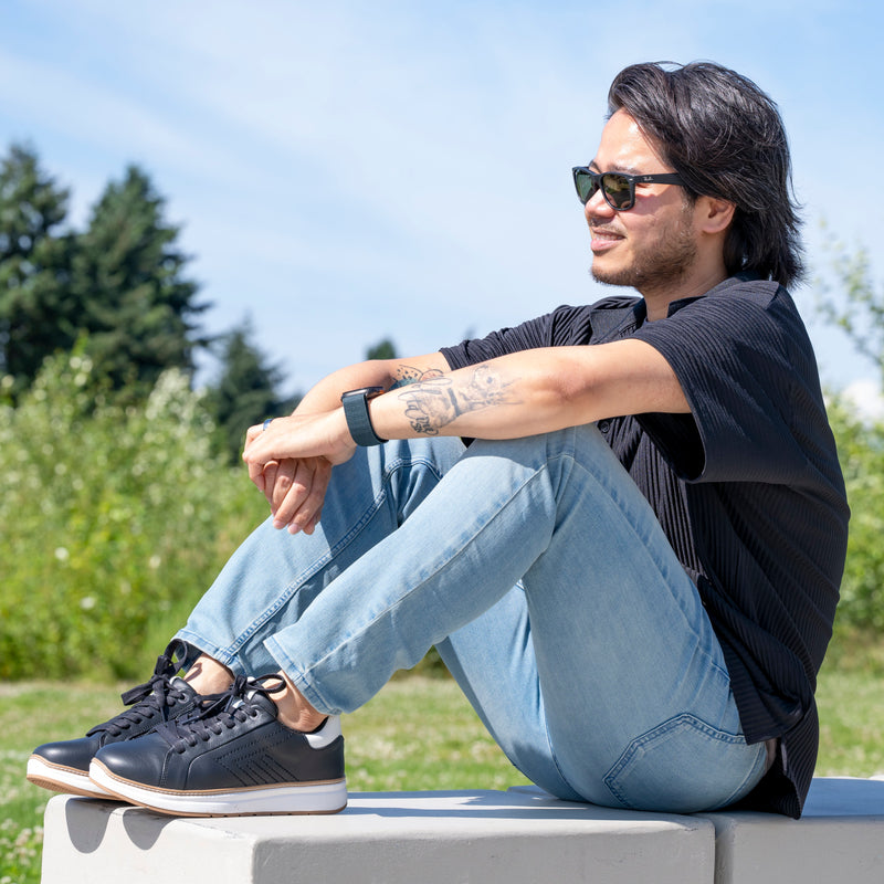 A man with long dark hair, sunglasses, a black shirt, light blue jeans, and Men's Black Leather BILLY Comfort Luxe dress sneakers by BILLY Footwear sits on a concrete bench outdoors with greenery in the background.