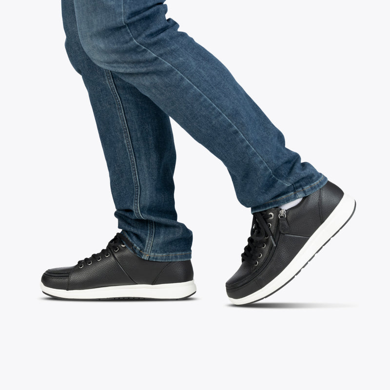 A person in blue jeans and Men's Black Leather BILLY Comfort Plush shoes by BILLY Footwear with white soles steps forward against a plain light background.