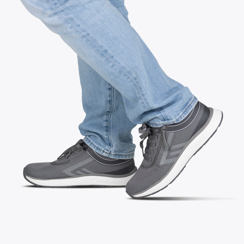 A person in light blue jeans steps forward on a white background, showcasing only the lower legs and feet in Men's Charcoal BILLY Sport Inclusion sneakers by BILLY Footwear, highlighting the inclusive design and style.