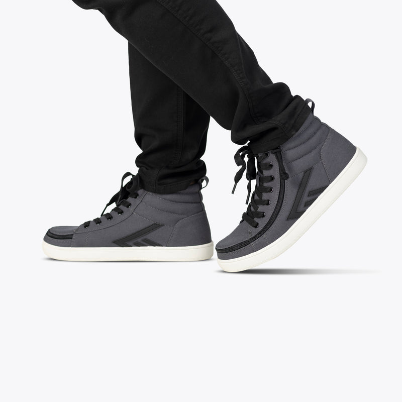 Wearing black jeans and BILLY Footwear’s Men's Charcoal/Black BILLY CS High sneakers with Full Wrap Zipper, this person walks against a plain light background.