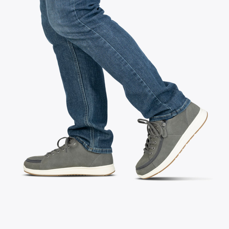 Wearing Men's Charcoal Suede BILLY Comfort Plush sneakers by BILLY Footwear, a person is shown mid-step against a plain white background, displaying only their lower legs and feet. The shoes feature a signature wraparound zipper.