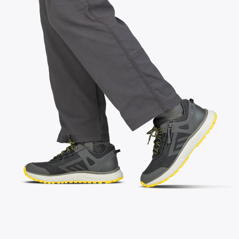 A person wears Men's Charcoal/Yellow BILLY Inclusion Trail sneakers from BILLY Footwear, featuring yellow soles and accents, paired with dark gray pants. Only their lower legs and feet are visible against a light background.