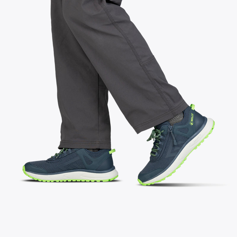 A person in dark gray pants wears BILLY Footwear's Men's Navy/Green BILLY Inclusion Trail—navy athletic slip-on hiking shoes with neon green accents—walking against a light gray background, with only lower legs and feet visible.