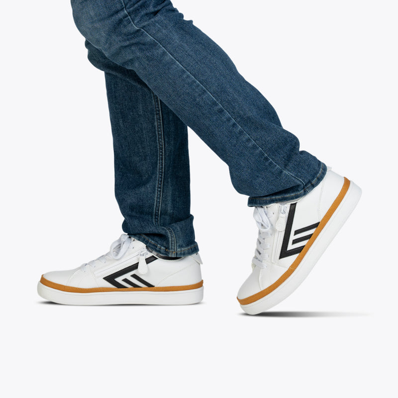 A person wears blue jeans and BILLY Footwear's Men's White/Black BILLY CS Low sneakers with black stripes and tan soles, walking against a plain light background. Only their lower legs and feet are shown.