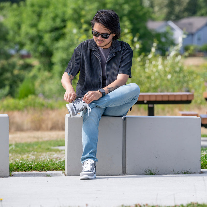 A man in sunglasses and casual clothes sits outdoors on a concrete bench, tying his BILLY Footwear Men's White/Grey/Black Comfort DZ High sneakers. Green trees, grass, and a blurred view of houses complete the relaxed scene.