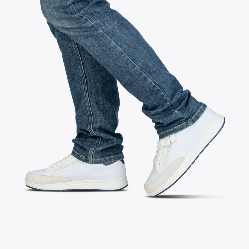 A person in blue jeans is walking, shown from the knees down against a plain light background, wearing Men’s White/Navy BILLY Comfort Court shoes by BILLY Footwear.