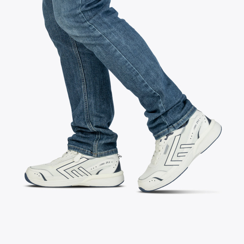 A person is shown walking in blue jeans and BILLY Footwear's Men's White/Navy BILLY Limitless 253 shoes, featuring easy-on/easy-off design, with only their lower legs visible against a plain light background.