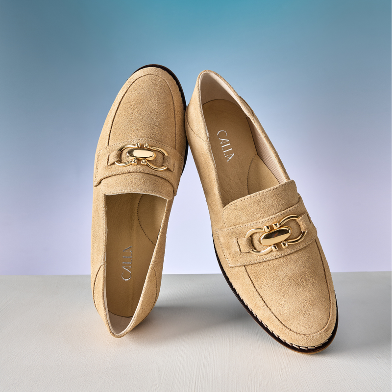 Pair of tan loafers with a gold buckle on a light gray surface and blue gradient background