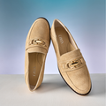 Pair of tan loafers with a gold buckle on a light gray surface and blue gradient background