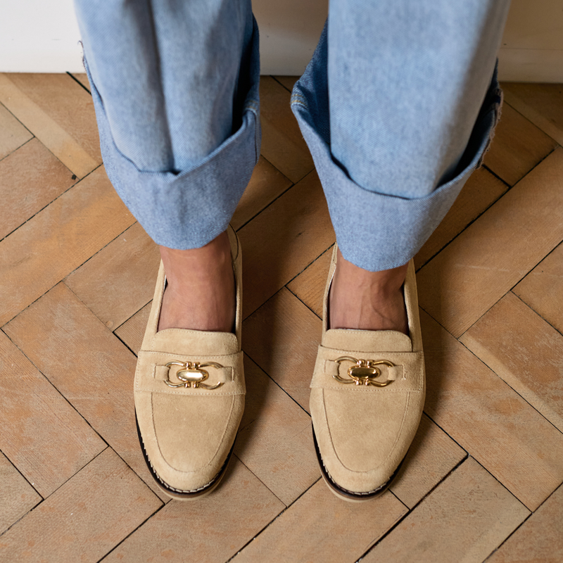 Beige loafers with gold buckles worn with light blue jeans on a wooden floor.