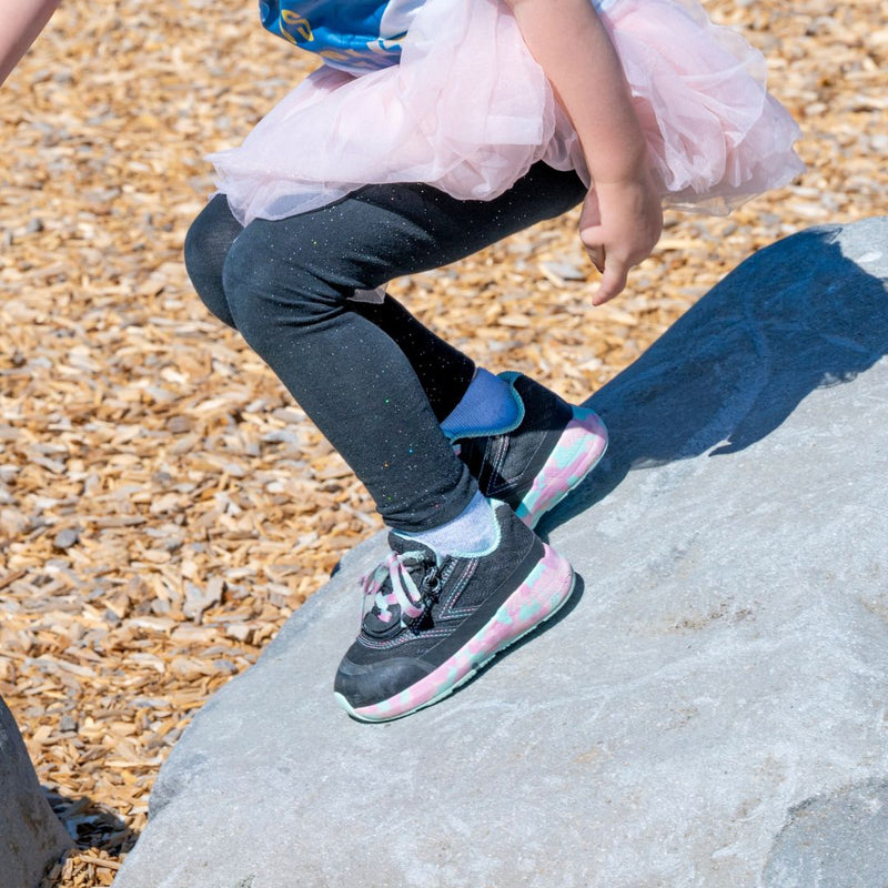 A child in black leggings, a pink tutu, and BILLY Footwear's Midnight Mermaid BILLY Goat sneakers with pastel soles climbs a large gray rock at a wood chip-covered playground.