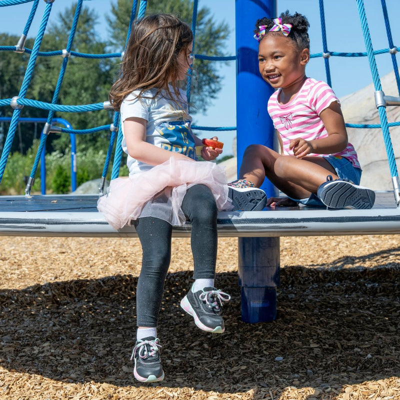 Two young girls smile and chat on a playground climber. One wears a tutu, leggings, and BILLY Footwear's Midnight Mermaid BILLY Goat shoes, designed to be AFO-friendly. The other sports a striped shirt with bows in her hair. Wood chips cover the ground below.
