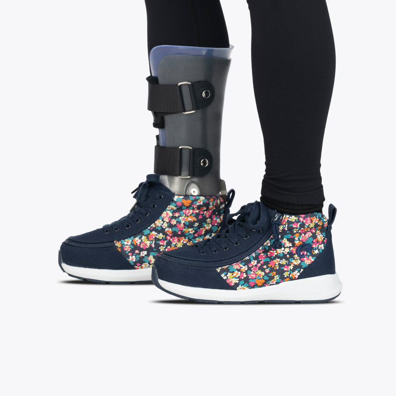 A person wears black leggings with a gray orthopedic brace over one leg and shoe. They sport Navy Floral BILLY Goat Classic High sneakers by BILLY Footwear. The background is plain white.