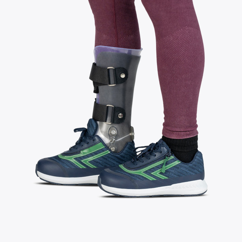 A person in purple leggings and a gray orthopedic brace with black straps wears Navy/Green BILLY Goat sneakers by BILLY Footwear, demonstrating easy-on/off AFO-friendly shoes.