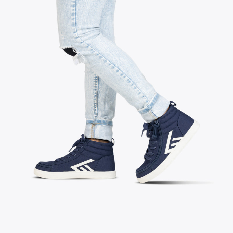 Wearing light-wash ripped skinny jeans and Navy/White BILLY CS High sneakers by BILLY Footwear with white soles and arrow designs, a person steps forward against a white background.