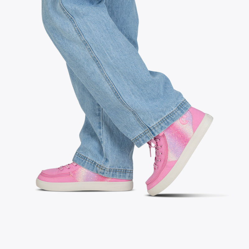 A person in light blue jeans and Pink Glitz BILLY Classic Lace High sneakers from BILLY Footwear, featuring white soles, sparkly gradient accents, and a wraparound zipper, is walking on a plain white background. The focus is on their lower legs and feet.