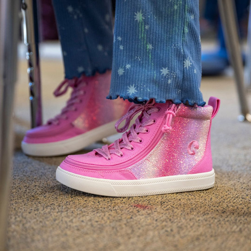 Close-up of a person wearing Pink Glitz BILLY Classic Lace High sneakers from BILLY Footwear, featuring wraparound zipper and white soles, standing on carpet. They have blue star-patterned pants with ruffled hems.