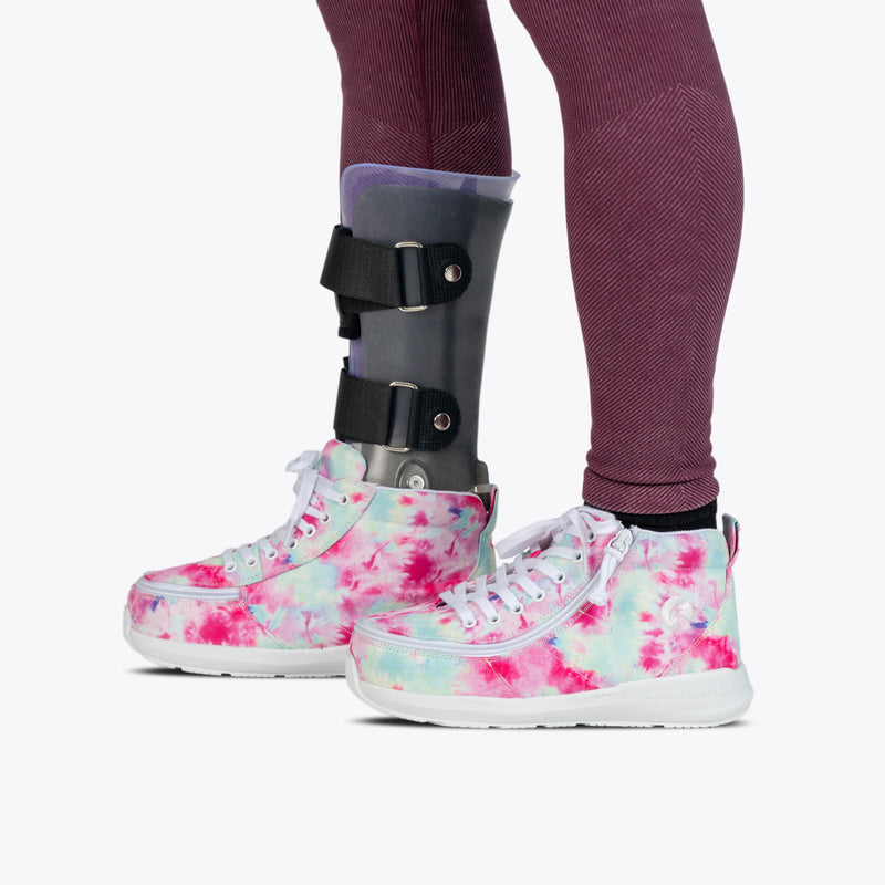 A person wearing maroon leggings and Pink Watercolor BILLY Goat Classic High adaptive footwear by BILLY Footwear, along with a black and gray orthopedic leg brace on their left leg.