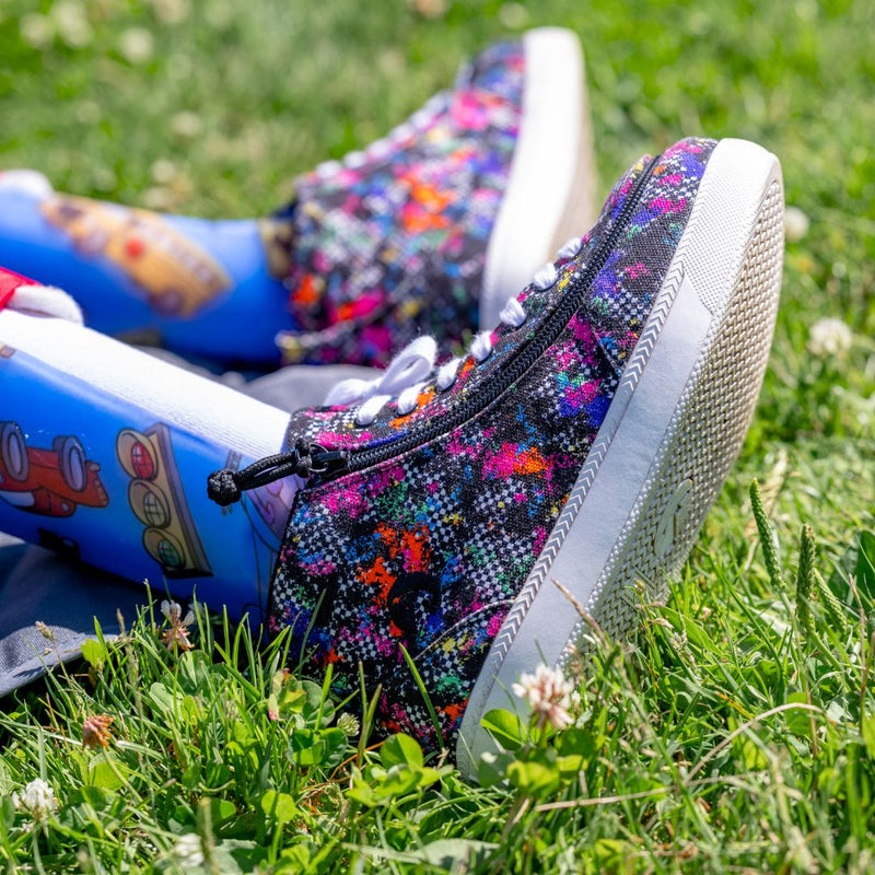 Close-up of a person with colorful prosthetic legs featuring cartoon designs, wearing BILLY Footwear Rainbow Sk8ter BILLY Classic D|R II High sneakers, sitting on grass with white clover flowers against a blue background.