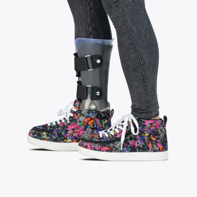A person wearing dark jeans and Rainbow Sk8ter BILLY Classic D|R II High shoes from BILLY Footwear, with a visible lower leg prosthesis on their right leg secured by black straps.