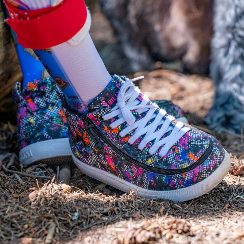 A close-up shows the Rainbow Sk8ter BILLY Classic D|R II High kids' orthopedic shoes by BILLY Footwear, featuring colorful patterns, white laces, and a side zipper. The shoes are worn on a forest floor with part of a prosthetic leg visible.