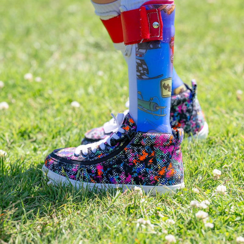 A child wearing colorful Rainbow Sk8ter BILLY Classic D|R II High shoes by BILLY Footwear stands on grass. One leg has a prosthetic with cartoon designs and a red knee attachment, surrounded by white clover flowers on the lawn.