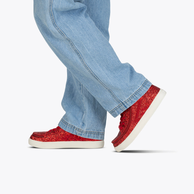 A person in light blue jeans and Ruby Red Glitz BILLY CS High sneakers by BILLY Footwear with white soles walks, one foot slightly raised, against a plain white background.