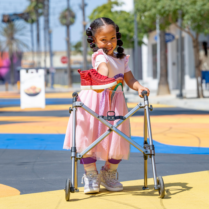 A young girl in a pink dress beams with confidence while using a walker outdoors on a sunny, palm-lined sidewalk, wearing shiny Ruby Red Glitz BILLY CS High sneakers from BILLY Footwear.