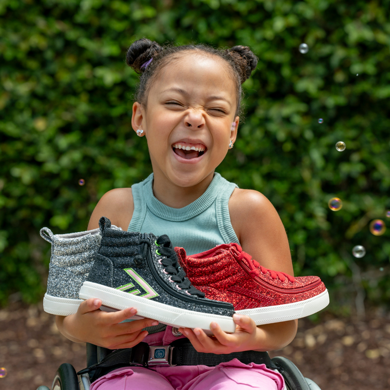 A smiling young girl in a sleeveless top and pink pants holds up black zipper shoes and BILLY Footwear's Ruby Red Glitz BILLY CS High tops outdoors, with green foliage and bubbles in the background.