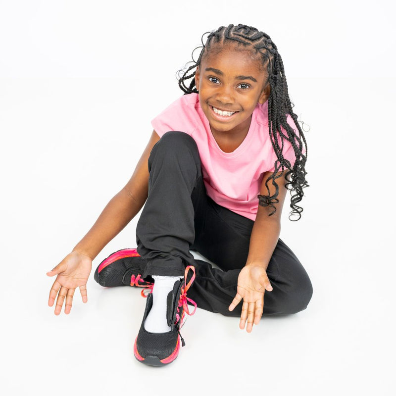 A smiling young girl with braided hair, pink shirt, and black pants sits on the floor in SALE - Black/Fuchsia Fire BILLY Sport Inclusion shoes by BILLY Footwear, one leg bent, hands open, looking up at the camera.